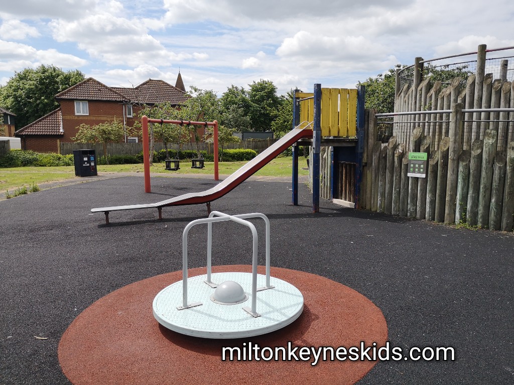 A small children's roundabout in front of a slide attacked to a wooden frame, with a swing behind the frame